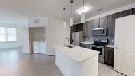 Located at 3420 Legacy Eagle Dr in Mount Pleasant, South Carolina, this single-floor residence offers a thoughtfully designed layout with contemporary finishes throughout. The open-concept kitchen serves as the heart of the home, anchored by a large center island with a striking quartz countertop, an undermount stainless steel sink, and elegant pendant lighting overhead. Dark wood-tone cabinetry with a textured finish lines the walls, complemented by a gray subway tile backsplash and a built-in wine rack for added character. Stainless steel appliances — including a French door refrigerator, electric range, microwave, and dishwasher — complete the space with a polished, modern aesthetic. Light wood-look flooring flows seamlessly throughout the home, enhancing the bright, airy atmosphere created by an abundance of natural light from multiple windows. The living areas are open and spacious, with ceiling fans and track lighting providing functional overhead illumination. A dedicated laundry closet houses a full-size washer and dryer, adding everyday convenience. The bathroom features a dual-sink vanity with a quartz countertop, dark wood-tone cabinetry with brushed nickel hardware, and a large framed mirror above backlit sconces. Bedrooms are generously sized with ample natural light and direct access to additional storage areas, making this a well-appointed and move-in-ready home.