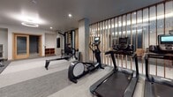 This modern fitness center is thoughtfully designed to accommodate a wide range of workout styles and fitness goals. The space is organized into distinct functional zones, creating an efficient and intuitive layout for users. The interior features a neutral color palette of grays and whites, accented by warm wood tones from slatted divider panels and door frames, lending the space a contemporary yet approachable aesthetic. Exposed concrete columns add an industrial character that complements the overall design.

The cardio zone is well-appointed with multiple treadmills, an elliptical trainer, and a rowing machine, while a wall-mounted flat-screen television enhances the experience. A dedicated strength area houses a cable machine with an adjustable bench, a multi-functional training wall with a stowable punching bag, and a full rack of dumbbells. Mirrored walls expand the sense of space and allow users to monitor their form. Rubber flooring in key workout zones provides durability and safety. A built-in water station with dual drinking fountains and a bottle-filling option ensures easy hydration. A separate room with glass-paneled doors offers additional private workout space, completing this comprehensive fitness facility.