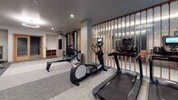 This modern fitness center is thoughtfully designed to accommodate a wide range of workout styles and fitness goals. The space is organized into distinct functional zones, creating an efficient and intuitive layout for users. The interior features a neutral color palette of grays and whites, accented by warm wood tones from slatted divider panels and door frames, lending the space a contemporary yet approachable aesthetic. Exposed concrete columns add an industrial character that complements the overall design.

The cardio zone is well-appointed with multiple treadmills, an elliptical trainer, and a rowing machine, while a wall-mounted flat-screen television enhances the experience. A dedicated strength area houses a cable machine with an adjustable bench, a multi-functional training wall with a stowable punching bag, and a full rack of dumbbells. Mirrored walls expand the sense of space and allow users to monitor their form. Rubber flooring in key workout zones provides durability and safety. A built-in water station with dual drinking fountains and a bottle-filling option ensures easy hydration. A separate room with glass-paneled doors offers additional private workout space, completing this comprehensive fitness facility.