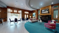 This elegantly designed single-floor space combines contemporary style with warm, inviting materials to create a refined communal environment. Rich wood paneling lines the walls throughout, providing a cohesive backdrop that contrasts beautifully with the bold teal accents found in built-in niches, area rugs, and decorative furnishings.

The main lounge area is a standout, anchored by a dramatic floor-to-ceiling black tile fireplace flanked by wood-framed display niches housing tall ceramic vases. A wall-mounted flat-screen television sits above the fireplace, while a large circular teal area rug defines the seating arrangement below a sweeping curved ceiling detail with recessed lighting. Plush sofas, vibrant magenta accent chairs, and a glass-top coffee table complete the sophisticated arrangement.

Adjacent to the lounge, a dedicated bar and kitchenette features a wraparound black granite countertop, woven-base bar stools, open shelving, globe pendant lighting, and full cabinetry — ideal for hosting and entertaining. A glass-top dining table with navy upholstered chairs sits nearby, bathed in natural light pouring through oversized windows and French doors that frame picturesque views of an outdoor pool area. Artwork, track lighting, and light hardwood floors round out this polished, resort-inspired space.
