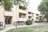 This single-floor covered carport is situated within a well-established residential complex surrounded by mature trees and open green spaces. The carport itself features a sturdy overhead structure with a flat ceiling and tan-painted panel walls on either side, providing shelter and protection from the elements. The concrete floor spans the length of the covered area, offering straightforward vehicle access from the adjacent roadway.

The exterior setting is characterized by a spacious lawn with mature deciduous trees that provide natural shade throughout the grounds. A concrete walkway connects the carport area to the surrounding buildings and common spaces, making pedestrian navigation convenient. The neighboring multi-story residential buildings share the same warm tan exterior siding, creating a cohesive visual aesthetic across the complex.

Views from within the carport look out onto the landscaped grounds, where additional trees, pathway lighting, and open grassy areas are visible. The complex appears to have multiple structures and shared outdoor common areas, giving the overall environment a community-oriented feel. The combination of covered parking, established greenery, and accessible walkways makes this carport a functional and well-integrated component of the broader residential grounds.