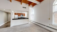 This well-appointed one-bedroom unit showcases a distinctive architectural character defined by soaring ceilings with warm, exposed wooden beams visible throughout the main living areas and bedroom. The open-concept kitchen and living room combination creates a spacious, functional layout anchored by an L-shaped peninsula with dark granite countertops, rich medium-toned wood cabinetry, and a full suite of stainless steel appliances including a refrigerator, electric range, and built-in microwave. Recessed lighting illuminates the kitchen, while a ceiling fan adds comfort to the living area. Elegant arched windows are a defining architectural feature, flooding the living room and bedroom with natural light and offering views of surrounding brick exteriors. The bedroom, accessed via a carpeted hallway with a double-door closet, continues the high-ceiling aesthetic with exposed beams overhead and its own arched window, creating an airy, open feel. Soft wall-to-wall carpeting runs through the living and sleeping areas, while the hallway transitions to rich hardwood flooring. Baseboard heating units line the perimeter walls throughout. The bathroom features a bold red vanity and louvered closet doors, adding a touch of contrast to the otherwise neutral palette.