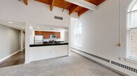 This well-appointed one-bedroom unit showcases a distinctive architectural character defined by soaring ceilings with warm, exposed wooden beams visible throughout the main living areas and bedroom. The open-concept kitchen and living room combination creates a spacious, functional layout anchored by an L-shaped peninsula with dark granite countertops, rich medium-toned wood cabinetry, and a full suite of stainless steel appliances including a refrigerator, electric range, and built-in microwave. Recessed lighting illuminates the kitchen, while a ceiling fan adds comfort to the living area. Elegant arched windows are a defining architectural feature, flooding the living room and bedroom with natural light and offering views of surrounding brick exteriors. The bedroom, accessed via a carpeted hallway with a double-door closet, continues the high-ceiling aesthetic with exposed beams overhead and its own arched window, creating an airy, open feel. Soft wall-to-wall carpeting runs through the living and sleeping areas, while the hallway transitions to rich hardwood flooring. Baseboard heating units line the perimeter walls throughout. The bathroom features a bold red vanity and louvered closet doors, adding a touch of contrast to the otherwise neutral palette.