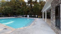 Located at 5533 Houston Rd in Macon, Georgia, this single-level community pool and recreational area offers a well-appointed outdoor retreat for residents. The centerpiece of the space is a generously sized swimming pool with clear blue water, tiled coping, and built-in entry steps complete with a handrail for easy access. A covered colonnade with white columns runs alongside the pool, providing a shaded walkway and enhancing the overall elegance of the setting.

The pool is enclosed by a black iron safety fence, ensuring security while maintaining an open, airy feel. Lush mature trees and natural Georgia greenery surround the perimeter, creating a serene, wooded backdrop that enhances the sense of privacy and tranquility.

Adjacent to the pool is an expansive concrete patio deck furnished with black wrought-iron lounge chairs, a round dining table, and a large navy blue market umbrella — ideal for outdoor relaxation and socializing. The accompanying clubhouse building features a striking natural stone exterior with varied earth-tone masonry, black decorative shutters, and large grid-pane windows that offer views into what appears to be an interior pool or recreation room. The combination of durable stone architecture, thoughtful outdoor furnishings, and mature landscaping makes this community amenity space both functional and visually appealing.
