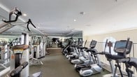 This spacious fitness center is thoughtfully designed to accommodate a full range of workout routines in a clean, professional environment. The single-floor layout is organized into distinct zones, separating cardio equipment from strength training areas for efficient use of the space. A robust lineup of True-brand treadmills and elliptical trainers lines one side of the room, while StairMaster steppers and additional cardio machines round out the aerobic offerings. The strength training section features a comprehensive array of cable machines and resistance equipment by Hoist and Cybex, suitable for full-body conditioning. Floor-to-ceiling mirrors span multiple walls, amplifying the sense of openness and allowing users to monitor their form throughout their workouts. The flooring is durable rubber gym matting, providing both comfort and safety. Recessed lighting illuminates the space evenly from a tray ceiling, while large arched windows along one wall allow natural light to filter in and offer views of the surrounding greenery. An iron railing along an open-sided section adds an architectural touch. The overall ambiance is bright, clean, and motivating, reflecting a well-maintained facility designed to support a serious fitness lifestyle.