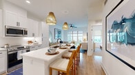 This well-designed single-floor unit offers a cohesive coastal-modern aesthetic across its thoughtfully arranged rooms. The open-plan kitchen and living area forms the heart of the space, unified by light hardwood-style flooring and crisp white walls. The kitchen is appointed with floor-to-ceiling white shaker cabinetry, black hardware accents, stainless steel appliances including a range, microwave, and refrigerator, and a white quartz island with an undermount sink. Woven gold pendant lights hang above the island, reinforcing the coastal character. Seating at the island connects the kitchen seamlessly to the living area, which features large windows framing greenery views and a ceiling fan overhead. Natural light fills the room, creating an airy, relaxed atmosphere.

The bedroom is a calm, well-proportioned retreat with a window, carpet flooring, and ample wall space. It connects directly to the full bathroom, which features a floating white vanity with a backlit mirror, black fixtures, light wood-tone flooring, and a glass-enclosed shower. A dedicated closet completes the private quarters. The consistent use of white tones, natural textures, and navy blue accents throughout gives the entire unit a cohesive, resort-inspired feel.