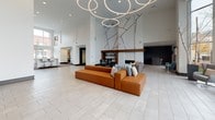 This single-floor clubhouse is designed with a contemporary aesthetic that balances open communal spaces with distinct functional areas. The grand entrance lobby immediately commands attention with soaring double-height ceilings, large-format tile flooring, and a striking circular chandelier installation overhead. An abstract geometric wall mural serves as a bold focal point, while a built-in fireplace and modular seating in warm cognac tones anchor the space with comfort and style.

Adjacent to the lobby, a well-appointed kitchen features rich wood cabinetry, granite countertops, a white subway tile backsplash, floating wood shelves, and stainless steel appliances, including a microwave and dishwasher. The kitchen opens to the main lounge area, promoting a seamless flow throughout the space.

A dedicated lounge area offers a mounted flat-screen television, a linear gas fireplace with wood accent surround, and comfortable seating arranged over patterned carpet tiles. Direct glass door access leads to a sunlit solarium-style sitting area lined with floor-to-ceiling windows overlooking the outdoor pool, furnished with wingback chairs and ornate accent tables. A separate game room accommodates a full-size pool table alongside additional seating, with pool views through large windows.
