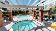 This outdoor amenity space is thoughtfully designed to offer residents a comprehensive recreational experience. At its center is a generously sized swimming pool with crystal-clear water, surrounded by an expansive concrete deck lined with lounge chairs and patio umbrellas, ideal for sunbathing and relaxation. Adjacent to the pool sits a circular hot tub, partially sheltered beneath a white pergola structure supported by decorative brick columns, which provides a shaded transition zone between the various outdoor areas.

The covered patio sections feature comfortable seating arrangements, including cushioned sofas, lounge chairs, and accent tables, creating inviting spaces for both casual conversation and unwinding. Grilling stations are conveniently integrated into the covered areas, enhancing the space's functionality for outdoor entertaining. Colorful accent furniture and ornamental grasses add visual interest throughout.

The architecture of the surrounding multi-story buildings incorporates a combination of brick, painted siding in warm earth tones, and large windows, contributing a residential warmth to the setting. Wrought iron fencing defines the perimeter of the amenity area, while mature trees and manicured landscaping provide natural shade and a pleasant green backdrop. The space benefits from abundant natural light under open skies, making it a vibrant and welcoming outdoor destination.