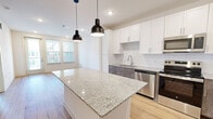 Located at 4997 Springdale Rd in Louisville, KY, this well-appointed one-bedroom, one-bathroom unit offers a thoughtfully designed layout with modern finishes throughout. The open-concept kitchen and living area serves as the heart of the home, anchored by a generous granite-topped island illuminated by a pair of sleek black pendant lights. The kitchen is outfitted with crisp white shaker cabinetry, contrasting wood-tone lower cabinets, and a full suite of stainless steel appliances, including a French door refrigerator, range, built-in microwave, and dishwasher. Light hardwood-style flooring runs throughout the main living areas, enhancing the bright, airy feel. The bedroom is a comfortable retreat featuring plush carpet, a ceiling fan, and dual windows that invite natural light. The bathroom is finished with a granite-topped vanity, wood-tone cabinetry, and direct access to a dedicated walk-in closet with wire shelving for ample organization. A conveniently located in-unit laundry area houses a full-size washer and dryer. Clean white walls and recessed lighting throughout give this unit a fresh, contemporary ambiance.
