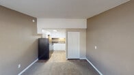 This single-floor unit offers a practical and functional layout designed to maximize the use of available space. The main living area is finished with neutral-toned carpet and warm beige walls, creating a comfortable and understated atmosphere. A window along one wall allows natural light to filter in, illuminating the space and providing an outdoor view. A wall-mounted HVAC unit ensures year-round climate control.

The kitchen is neatly tucked into an alcove off the living area and is well-appointed with crisp white shaker-style cabinetry extending from floor to ceiling, offering ample storage. Dark granite-style countertops provide a contrasting work surface, complemented by a stainless steel electric range with overhead ventilation, a stainless steel refrigerator, and a single-basin sink. The kitchen flooring transitions to light wood-look planks, adding a clean, distinct separation from the carpeted living space.

The bathroom features a white vanity with an integrated sink, a large mirror, chrome fixtures and towel bars, and textured neutral wall finishes. A shower/tub combination completes the space. A panel-style closet door in the main area provides convenient storage access. The textured ceiling is consistent throughout, lending a cohesive character to the overall unit.