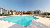 Located at 148 Wall St in Camden, South Carolina, this outdoor community amenity space offers a well-appointed resort-style environment designed for relaxation and social gatherings. The centerpiece is a generously sized swimming pool with crystal-clear blue water, accessible via stainless steel entry rails and surrounded by a broad concrete sun deck. The deck is furnished with lounge chairs and shaded by umbrellas, providing ample space for sunbathing and leisure under the open South Carolina sky. Tall, mature palm trees line the pool area, lending a tropical ambiance that complements the sunny climate. A white perimeter fence encloses the pool zone, offering both safety and a sense of privacy. Adjacent to the pool, a well-landscaped patio area features outdoor dining furniture, including a round table with a large blue umbrella and additional seating, alongside a raised bar area perfect for entertaining. Decorative planters with colorful seasonal blooms and ornamental grasses add vibrant touches throughout. The surrounding buildings feature neutral siding, pitched rooflines, and private balconies that overlook the amenity area. The overall atmosphere is inviting and community-oriented, offering residents a comfortable and attractive outdoor retreat.