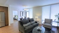This single-floor residence is thoughtfully designed with a clean, contemporary aesthetic that emphasizes comfort and functionality. A central hallway serves as the spine of the unit, connecting the various living spaces with white-trimmed doorways and smooth, light-toned hardwood flooring that extends throughout. Recessed lighting in the corridor provides a welcoming transition between rooms.

The living area is generously proportioned and bathed in natural light from large windows dressed with horizontal blinds. The space comfortably accommodates a plush, dark upholstered sofa paired with coordinating accent chairs, a glass-top coffee table, and a standing lamp, creating a relaxed yet polished atmosphere. A wall-mounted mirror and a small dining or accent table add depth and dimension to the room.

Visible through an open doorway, the bedroom continues the hardwood flooring and benefits from its own window, maintaining the unit's bright and airy character. The neutral color palette of warm whites and soft beige tones throughout creates a cohesive and calming ambiance, making the space feel inviting and well-balanced. The overall layout is efficient, ensuring each area flows naturally into the next.