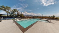 This expansive outdoor amenity space is designed for relaxation and recreation, centered around a generously sized swimming pool with crystal-clear turquoise water. The pool deck is finished in smooth concrete and lined with rows of modern white lounge chairs, providing ample space for sunbathing and leisure. A dedicated hot tub offers a separate hydrotherapy area adjacent to the main pool, set against the backdrop of a contemporary building with clean, modern architectural lines featuring a neutral palette of gray and white panels.

The space is further enhanced by a substantial pergola structure that spans a portion of the deck, providing shaded seating and gathering areas below. A private cabana with flowing white curtains offers an additional retreat for guests seeking a more secluded experience. Manicured green turf accents are incorporated throughout the deck, adding pops of color and softening the hardscape.

The entire pool area is enclosed by sleek black iron fencing, ensuring privacy while maintaining open sightlines. The surrounding landscape features mature trees that frame the space naturally. Most notably, the amenity area commands sweeping views of a dramatic mountain range, creating a striking scenic backdrop visible from nearly every angle of the pool deck. Open skies above complete the resort-like atmosphere of this outstanding outdoor space.