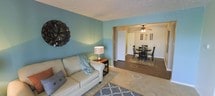 This single-floor space offers a comfortable and functional layout, with a living area that flows seamlessly into an adjacent dining room through an open doorway. The living area features soft carpeting, teal-painted walls, and a textured ceiling, creating a casual and welcoming ambiance. A side table with a decorative lamp adds a warm lighting element, complementing the neutral-toned sofa. A decorative wall accent adds a touch of personality to the space.

The transition into the dining area is defined by a change in flooring, shifting from carpet to hardwood, which adds visual contrast and delineation between the two spaces. The dining area is equipped with a ceiling fan that provides both lighting and air circulation. A round dining table surrounded by dark wooden chairs sits atop a patterned area rug, anchoring the room. A white door visible in the background suggests access to additional areas of the home. Together, the two spaces combine practicality with a relaxed aesthetic, making the layout well-suited for everyday living and casual entertaining.