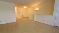 This single-floor space offers a comfortable and functional layout with a warm, inviting aesthetic. The main living area is generously proportioned, finished with soft, neutral-toned wall-to-wall carpeting that adds warmth underfoot. Smooth, cream-colored walls are accented by classic white chair rail molding that runs throughout, lending a traditional architectural character to the interior.

A standout feature of the main room is the decorative fireplace surround with a clean white mantel, serving as a natural focal point and adding charm to the space. Overhead, a centrally mounted ceiling light fixture provides ambient illumination, while a wall-mounted sconce adds supplemental lighting and a subtle decorative touch.

A floor vent is visible along the baseboard, indicating a built-in climate control system for year-round comfort. The layout flows openly, with a hallway visible in the background suggesting additional rooms or spaces beyond the main area. Clean white baseboards frame the perimeter of the room, complementing the overall neutral and well-maintained interior palette.

The space presents a clean, move-in-ready condition with consistent finishes throughout, making it a versatile environment well-suited for a variety of uses and furnishing styles.