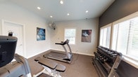 The Aura fitness center is a thoughtfully designed, single-floor amenity space built to support a well-rounded workout routine. The main exercise area features carpeted flooring that provides comfort and traction during workouts. Natural light filters in through multiple windows fitted with blinds, brightening the space alongside recessed ceiling lighting that ensures consistent illumination throughout the room. A wall-mounted industrial fan adds practical ventilation during intense sessions.

The cardio zone is equipped with a True treadmill and an upright stationary bike, offering residents reliable options for aerobic training. A dumbbell rack stocked with a full range of free weights lines one wall, while a weight bench provides space for strength-focused exercises. Sport-themed wall art adds a motivating visual energy to the room.

A distinctive feature of the fitness area is its interior window wall, which opens visually into an adjacent space — a dedicated business center. That connected area houses computer workstations and is accessible through a glass-paneled door, making it easy to transition between fitness and productivity. The overall design of the space is clean and functional, with neutral tones and a straightforward layout that keeps the focus on fitness.