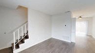 This two-story space offers a fresh, modern aesthetic defined by clean lines and a neutral color palette. The main level features durable hardwood-style flooring in a warm gray tone, providing a contemporary foundation that complements the crisp white walls and white baseboards throughout. The open layout of the main living area creates a sense of spaciousness, enhanced by smooth ceilings with a subtle texture and visible HVAC vents that speak to the functional infrastructure of the space. A staircase with white-painted railings and dark treads connects the two floors, adding a visual contrast that anchors the open layout. The walls are freshly finished, offering a move-in-ready feel with ample flexibility for personalization. Natural light filters into the space, contributing to the bright and airy ambiance. The overall condition of the space suggests recent updates or renovation, with attention to detail evident in the clean finishes and consistent design language carried throughout. This two-level layout provides a practical separation of living areas across floors, making it a versatile and functional space.