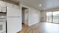 This single-floor unit offers a clean, modern aesthetic with a thoughtfully open layout that connects the living and kitchen areas seamlessly. Light-toned hardwood flooring runs throughout, lending warmth and continuity to every room.

The kitchen is well-appointed with white shaker-style cabinetry, ample upper and lower storage, light countertops, and a full suite of white appliances including a refrigerator, electric range, built-in microwave, and dishwasher. A single-basin sink with a contemporary pull-down faucet completes the functional workspace.

The open living area is bathed in natural light, enhanced by recessed ceiling lighting and a sliding glass door that leads to a private balcony with exterior views. A wall-mounted air conditioning unit provides climate control. The space offers generous room for a variety of furniture arrangements.

The bedroom is a comfortable, well-lit room featuring a window with a balcony view, a wall-mounted AC unit, and recessed lighting overhead. The bathroom is neatly finished with hardwood-style flooring, a sliding glass-door shower stall, and clean white fixtures. Overall, the unit presents a bright, move-in-ready environment with a cohesive and contemporary design sensibility.