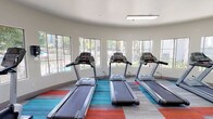 Located at 7131 Alvern St in Los Angeles, CA, this fitness center is a well-equipped, single-story facility designed to support a comprehensive cardio workout experience. The space features a distinctive curved wall lined with large windows that flood the interior with abundant natural light and offer views of the surrounding landscaped outdoor area, creating an inviting and energizing atmosphere.

The flooring throughout is a combination of neutral gray wood-look planks accented by vibrant multi-colored carpet tile insets in teal, red, and orange, adding a modern, dynamic visual element to the space. The ceilings are smooth and white, contributing to the open, airy feel of the room.

The fitness center is outfitted with Matrix brand cardio equipment, including three treadmills positioned along the curved window wall, multiple elliptical cross-trainers, and a recumbent stationary bike. A large floor-to-ceiling mirror along one wall visually expands the space and allows users to monitor their form during workouts. Glass doors mark the entrance, where posted fitness center rules are prominently displayed. Overhead lighting supplements the natural light, ensuring the space remains well-lit throughout the day.