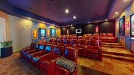 Located at 3311 Woodhill Ln in Fayetteville, NC, this single-level private home theater is a masterfully designed cinematic retreat that replicates the full commercial movie theater experience. The screening room is appointed with multiple rows of plush, red leather reclining seats arranged in a tiered, stadium-style layout, ensuring unobstructed sightlines from every position. Each seat features built-in cupholders, adding a practical touch to the luxurious setting. The room's deep jewel-toned color palette—rich blues, warm golds, and dark charcoal wall panels—creates an immersive, cinematic atmosphere. Illuminated movie poster displays with backlit frames line the walls, evoking the ambiance of a professional cinema lobby. Recessed ceiling lighting and strategically placed sconces provide layered, dimmable illumination throughout the space. The entry area features a dedicated lobby-style foyer complete with a large backlit "Coming Soon" movie poster display flanked by decorative planters, setting the tone before guests even enter the main screening room. A patterned tile and carpet flooring combination adds visual texture and further reinforces the theatrical aesthetic. An adjoining outdoor area with a pool is accessible from the space, rounding out this exceptional entertainment destination.