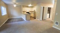 This single-floor residence offers a functional and open layout that seamlessly connects the living and kitchen areas into one cohesive space. The living room features neutral-toned carpet that adds warmth underfoot, while a ceiling fan overhead provides year-round comfort. A smooth transition leads into the kitchen, where hardwood-style flooring takes over, providing both practicality and visual contrast. The kitchen is well-equipped with a stainless steel refrigerator, a black electric range, and a built-in microwave or range hood above the stove. Cream-colored cabinetry lines the walls, offering ample storage, while a peninsula with a countertop overhang creates a natural divide between the kitchen and living area and provides additional prep or casual dining space. A double sink is conveniently positioned along the peninsula. The neutral, light gray walls throughout create a clean and airy atmosphere, complemented by consistent white trim and baseboards. A doorway visible from the main area hints at additional rooms beyond, suggesting a thoughtfully organized floor plan. The overall ambiance is bright, practical, and move-in ready.