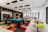 This single-floor amenity space is designed as a dynamic entertainment and social hub, blending modern design with playful elements. The open-plan layout is anchored by a full-size pool table set atop a bold, multicolored patchwork rug that adds a striking visual focal point to the room. Surrounding the rug, generous lounge seating is arranged around round coffee tables and ottoman poufs, creating an inviting gathering area. A wall-mounted television sits above a sleek fireplace, flanked by built-in shelving in a dark finish with gold accents, housing arcade machines and pinball machines that add a retro entertainment element. High-top tables with vibrant yellow chairs provide additional seating near a well-appointed kitchenette, which features white cabinetry, a clean countertop with an integrated sink, and a dramatic large-scale art mural. An array of sculptural pendant lights illuminates this prep area. Recessed linear lighting across the ceiling enhances the modern aesthetic throughout. Large floor-to-ceiling windows and glass doors flood the space with natural light and offer views of an outdoor terrace, seamlessly extending the entertaining area beyond the interior.