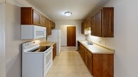 This 2-bedroom, 1-bathroom single-floor unit offers a straightforward and efficient layout designed for comfortable daily living. The galley-style kitchen is a standout feature, equipped with rich oak cabinetry spanning both upper and lower cabinets, a double-basin sink, light laminate countertops, white appliances including a range, refrigerator, microwave, and dishwasher, and tile flooring. The kitchen opens toward a living or dining area finished with carpet and a ceiling fan with light fixture overhead. Both bedrooms are carpeted and feature neutral wall tones with wood baseboard trim. The primary bedroom includes a sliding-door closet with a hanging rod, while the second bedroom benefits from a large window with vertical blinds that brings in natural light, complemented by a baseboard heater. The full bathroom is clean and functional, featuring a tub/shower combo, toilet, and tile flooring. Multiple hallways connect the rooms throughout the unit, contributing to an organized flow. Textured ceilings are present throughout the space. Overall, the unit presents a practical and well-organized interior with consistent warm-toned wood finishes tying the spaces together.