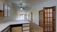 This single-floor space offers a thoughtfully open layout that connects the kitchen and living areas seamlessly. The kitchen features white cabinetry, a built-in dishwasher, and a peninsula countertop that flows naturally into the carpeted main living area. Vaulted ceilings with a ceiling fan add a sense of height and airiness throughout the shared space, while a wall-mounted air conditioning unit provides functional climate control.

The living area is generously proportioned, bathed in natural light from a sliding glass door that leads directly to a private wooden deck. From the deck, the view opens onto an expansive, park-like landscape with a well-maintained lawn and mature bare-branched trees spread across the grounds, offering a peaceful and scenic outdoor setting.

A distinctive wood-framed glass-panel door adds a classic architectural detail, providing separation between living spaces while maintaining visual openness. Warm wood trim throughout the unit complements the neutral carpet and light-toned walls, creating a cohesive and inviting interior. Additional rooms branch off from the main living space, offering privacy and versatility. Overall, the space combines practical design with natural surroundings for a comfortable and tranquil living environment.
