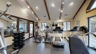 Located at 5350 Silverthorne Road in Westerville, Ohio, this fitness center offers a well-equipped and thoughtfully designed workout space on a single floor. The room is characterized by vaulted ceilings accented with rustic exposed wooden beams, adding warmth and architectural interest to the space. Track lighting and recessed ceiling fixtures ensure the area is brightly and evenly illuminated throughout.

The fitness floor features a selection of cardio machines, including treadmills, ellipticals, and a stair climber, arranged to allow for comfortable movement between equipment. A dedicated free weight station includes a full rack of dumbbells, catering to strength training needs. Wall-mounted mirrors expand the sense of space and allow users to monitor their form, while mounted television screens provide entertainment during workouts.

Large windows along the walls welcome natural light and offer views of the surrounding outdoor scenery, creating an open and energizing atmosphere. The durable flooring is well-suited for high-traffic fitness use. A glass-paneled door connects to an adjacent area, enhancing the overall flow of the facility. The space strikes a balance between functional fitness infrastructure and a clean, welcoming aesthetic.