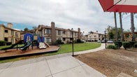 Located at 2130 S Santa Fe in Vista, this well-appointed outdoor community space serves as a central gathering hub within a multi-building residential complex. The grounds are thoughtfully laid out, offering a variety of amenities designed to accommodate residents of all ages. A standout feature is the vibrant, modern playground equipped with a multi-level play structure featuring climbing elements, slides, spiral climbers, and a wood chip safety surface — ideal for active play. Adjacent to the playground, a spacious concrete patio provides multiple picnic tables shaded by bold red-and-white striped umbrellas, creating a comfortable outdoor dining and socializing area. Lush, manicured green lawns stretch across the central courtyard, framed by mature palm trees and native landscaping that add a Southern California character to the setting. Decorative lamp posts and winding pathways connect the various areas of the complex, contributing to a welcoming and navigable environment. A wood privacy fence borders portions of the property, offering a sense of enclosure and security. The surrounding two-story stucco buildings, each featuring chimneys and exterior staircases, complete the community aesthetic. The overall ambiance is open, family-friendly, and well-maintained, providing a pleasant outdoor retreat for residents.