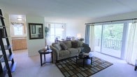 This single-floor residence features an open-concept layout that seamlessly connects the living and dining areas, creating a spacious and airy atmosphere. The living area is anchored by plush, neutral-toned seating arranged around a geometric patterned area rug, complemented by a round side table and a pair of low-profile coffee tables. Decorative throw pillows and a framed wall mirror add character to the space. Natural light flows generously through large windows and a sliding glass door that opens onto a private balcony with lush tree views, enhancing the connection between indoor and outdoor spaces. Sheer curtains frame the sliding door, softening the natural light while maintaining openness. The dining area, visible from the living space, features a pendant light fixture above a dining table with seating for several guests. Light-toned walls and neutral carpeting throughout contribute to a bright, cohesive aesthetic. A decorative indoor plant adds a refreshing natural element, while recessed architectural details in the ceiling provide subtle visual interest overhead.