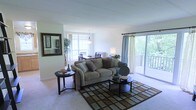 This single-floor residence features an open-concept layout that seamlessly connects the living and dining areas, creating a spacious and airy atmosphere. The living area is anchored by plush, neutral-toned seating arranged around a geometric patterned area rug, complemented by a round side table and a pair of low-profile coffee tables. Decorative throw pillows and a framed wall mirror add character to the space. Natural light flows generously through large windows and a sliding glass door that opens onto a private balcony with lush tree views, enhancing the connection between indoor and outdoor spaces. Sheer curtains frame the sliding door, softening the natural light while maintaining openness. The dining area, visible from the living space, features a pendant light fixture above a dining table with seating for several guests. Light-toned walls and neutral carpeting throughout contribute to a bright, cohesive aesthetic. A decorative indoor plant adds a refreshing natural element, while recessed architectural details in the ceiling provide subtle visual interest overhead.