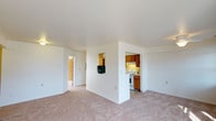 This single-floor space offers a clean, practical layout centered around a spacious, open living area finished with light beige wall-to-wall carpeting and crisp white walls and ceilings. The living area is bright and airy, providing a comfortable, flexible space suitable for various furniture arrangements.

A partial dividing wall separates the living area from the kitchen, which is accessible through a dedicated opening. The kitchen features warm, honey-toned wood cabinetry providing ample storage, a white gas range, and under-cabinet lighting that adds a functional glow to the workspace. The layout is efficient and well-organized, making practical use of the available space.

A hallway visible from the main living area leads to additional rooms beyond, suggesting a thoughtful flow throughout the unit. The interior is uniformly finished with smooth white ceilings and neutral-toned walls, lending a cohesive and clean aesthetic throughout. Overall, the space presents a straightforward, move-in-ready interior with a calm, neutral palette suited to a range of personal styles.