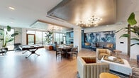 Located at 350 Carraway Crossing in Chapel Hill, NC, this sophisticated single-floor community lounge offers a generously proportioned, open-plan living space designed for both relaxation and social interaction. Light hardwood flooring runs throughout, anchoring the contemporary aesthetic and providing a warm contrast to the varied seating arrangements.

The space is organized into several distinct functional zones. A prominent seating area features a rich brown leather sectional sofa paired with tan channel-tufted armchairs, centered around a woven ottoman coffee table. A dedicated dining area offers a round table with upholstered chairs, ideal for casual gatherings. A billiards table provides a focal point for recreation near expansive floor-to-ceiling windows that flood the space with natural light and offer views of the surrounding landscape.

A striking floor-to-ceiling fireplace wall clad in large-format gray tile commands attention, complete with a linear gas fireplace, a natural wood mantel, and a wall-mounted television above. An adjacent lounge nook showcases dramatic blue marbled wall art and intimate seating. Tray ceilings with metallic gold finishes and Sputnik-style chandeliers add an elevated, upscale character throughout. Tall tropical plants punctuate the interior, enhancing the vibrant, welcoming ambiance of this well-appointed communal space.