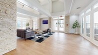 Located at 8340 N Tryon St in Charlotte, NC, this single-floor amenity space offers a generously sized, open-concept layout designed to serve as a multi-functional gathering and recreation hub. The interior is defined by soaring coffered ceilings, warm light wood flooring, and an abundance of natural light streaming through large windows and glass-paneled doors that open to an outdoor area.

The main lounge area is anchored by stacked stone columns that add a striking architectural contrast to the bright white walls and crown molding. Comfortable seating, a wall-mounted display, and a checkered area rug create an inviting social environment. Adjacent to the lounge, a fitness room is accessible through sleek black-framed glass doors, housing a visible array of exercise equipment and free weights.

The kitchen is thoughtfully designed with a two-tone cabinetry scheme — dark navy lower cabinets paired with crisp white uppers — complemented by quartz countertops, a subway tile backsplash, and a central island with bar seating. Industrial-style pendant lights provide focused illumination, while open shelving adds a decorative, functional touch. The overall ambiance of the space is clean, modern, and welcoming, blending classic architectural elements with contemporary design details.