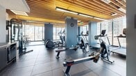 This fitness center offers a spacious, single-floor layout designed to accommodate a comprehensive range of workout equipment and activities. The space is defined by its striking architectural ceiling — a slatted wood panel design that adds warmth and texture, complemented by integrated linear lighting that ensures the area is well-lit throughout.

Floor-to-ceiling windows line the perimeter, flooding the interior with natural light and providing sweeping panoramic views of the surrounding urban skyline. The rubber-tiled flooring throughout provides both durability and safety for all types of exercise.

The facility is organized into distinct workout zones. The cardio area is well-stocked with treadmills, elliptical trainers, stationary bikes, and rowing machines, offering users a wide variety of aerobic exercise options. A free weights and strength training section features benches and additional resistance equipment. Blue-painted structural columns punctuate the space and subtly define each zone.

Adding a distinctive artistic touch, bold botanical murals — featuring large-scale floral illustrations in vivid pinks and greens — are displayed on accent walls, creating a visually dynamic atmosphere that balances the industrial aesthetic of the equipment. The overall ambiance is modern, energizing, and thoughtfully designed to inspire an effective workout experience.