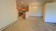 This single-floor residence offers a practical and welcoming open-concept layout that seamlessly connects the kitchen, dining, and living areas. The kitchen is well-appointed with warm honey-toned wood cabinetry, a white refrigerator, and a white electric range, all complemented by recessed lighting that ensures bright, even illumination throughout the cooking space. A peninsula provides additional counter space and serves as a natural divider between the kitchen and the adjoining living and dining areas. The dining area is distinguished by an elegant chandelier that adds a refined touch to the open floor plan. Soft, neutral-toned carpeting extends across the living area, creating a comfortable and cohesive atmosphere. Baseboard heating runs along the perimeter, offering efficient climate control throughout the space. The overall design balances functionality with a clean, understated aesthetic, making it a practical and comfortable living environment. The open sightlines between rooms enhance the sense of spaciousness, allowing natural flow and connectivity across the main living spaces.