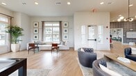Located at 1120 Corner Wind Ln in Columbia, SC, this single-floor community space offers a thoughtfully designed open-concept layout that seamlessly blends a fully equipped kitchen with a generous living and recreational area. The kitchen is a standout feature, showcasing a large waterfall-edge island with seating, dark gray cabinetry accented with gold hardware, white quartz countertops, a subway tile backsplash in soft blue-gray tones, open wood shelving, stainless steel appliances, and bold black pendant lighting overhead. The living area is expansive and versatile, featuring light hardwood-style flooring throughout, high ceilings with recessed lighting, and multiple seating arrangements including tufted leather chairs and sofas. A pool table and shuffleboard table add a recreational dimension to the space. Large windows with neutral drapery line the walls, flooding the interior with natural light and offering views of the surrounding community. A dedicated nook features a built-in counter with bar seating and an eye-catching geometric accent wall, providing an ideal spot for casual work or games. French doors with glass panels open to outdoor areas, reinforcing the bright, airy ambiance. The overall aesthetic is modern and inviting, combining clean lines with warm, welcoming touches.