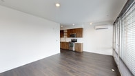 This single-floor space showcases a clean, contemporary design defined by an open-plan layout that seamlessly integrates the living area and kitchen. Dark hardwood flooring runs throughout, providing a warm contrast against the bright white walls and smooth white ceiling, which is fitted with recessed lighting and a track lighting system that ensures the space is well-illuminated throughout.

The kitchen is positioned along the back wall and features rich, wood-grain cabinetry with a distinctive horizontal grain pattern, offering ample upper and lower storage. Open shelving adds a modern touch above the countertops, while the light-colored counters provide a clean, functional workspace. Stainless steel appliances, including a freestanding electric range and an over-the-range microwave, complete the kitchen with a polished, modern aesthetic. A single-basin sink with a modern faucet is integrated into the countertop.

Climate control is handled by a wall-mounted mini-split air conditioning unit positioned along the side wall. The open living area is generously proportioned, offering a versatile and airy feel that lends itself well to a variety of furniture arrangements. The overall ambiance is modern, minimal, and functional.
