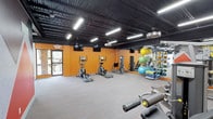 This fitness center offers a spacious, open layout designed to accommodate a wide range of workout activities. The room features bold orange accent walls that create an energetic atmosphere, contrasting with the dark, exposed ceiling overhead. Industrial-style fluorescent lighting is suspended from above, ensuring the space is well-lit throughout.

The cardio area is equipped with multiple elliptical machines positioned along the floor, providing ample space between each unit for comfortable use. Mounted flat-screen televisions are positioned on the walls, allowing members to stay entertained during their workouts. Along one wall, a large mirror extends the visual depth of the room while supporting functional training activities.

A dedicated storage rack holds a variety of exercise balls in multiple sizes and colors, resistance equipment, and rolled mats, keeping accessories organized and accessible. Free weight equipment is visible in the foreground, indicating a strength training area within the same open space.

The flooring consists of low-pile carpet, offering both comfort and durability underfoot. A glass-paneled entry door provides access and allows natural light to filter into the space, contributing to the overall bright and motivating environment.