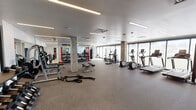 This well-appointed fitness center is designed to support a comprehensive range of workout routines across several distinct functional zones. The space is generously proportioned, with smooth white ceilings fitted with linear LED lighting that provides consistent, even illumination throughout. Concrete structural columns punctuate the open floor plan, adding an industrial character to the otherwise clean, contemporary aesthetic.

The cardio zone lines one side of the facility, featuring a row of treadmills and elliptical trainers positioned along floor-to-ceiling windows that invite ample natural light and exterior views. A separate area accommodates stationary and spin bikes, set behind elegantly frosted glass panels etched with a floral motif, offering a sense of privacy while maintaining openness.

The strength and free-weight section is equipped with barbells, dumbbells, weight benches, battle ropes, and a punch bag, all arranged around mirrored walls that visually expand the space. Red power racks add a bold contrast against the neutral palette.

A dedicated studio area features light hardwood flooring, full-length mirrors, and freestanding mirror panels, making it suitable for stretching, yoga, or personal training sessions. Rubber-floored zones provide safety and durability in high-traffic workout areas. The facility is accessed through glass-paneled doors, reinforcing the space's modern, transparent design language.