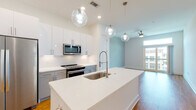 Located at 5126 Peachtree Blvd in Chamblee, Georgia, this single-floor residence offers a thoughtfully designed open-concept layout with modern finishes and an abundance of natural light. The kitchen serves as the heart of the home, anchored by a large center island with a white quartz countertop and an undermount stainless steel sink with a professional-style faucet. Crisp white upper cabinets are complemented by gray lower cabinets, while stainless steel appliances — including a refrigerator, range, and microwave — complete the contemporary look. Globe pendant lights add a stylish accent overhead. Hardwood flooring flows through the main living areas, creating a warm and cohesive aesthetic. The open living area benefits from direct access to a private balcony through sliding glass doors topped with a transom window, bringing in generous outdoor views and natural light. The bedroom features plush carpeting, a ceiling fan, decorative wall paneling, and a well-organized walk-in closet with built-in shelving. The bathroom is finished with a glass-enclosed walk-in shower, gray vanity cabinetry with brushed nickel hardware, and white quartz countertops. Clean lines, a soft neutral palette, and ceiling fans throughout contribute to the space's bright, modern, and comfortable ambiance.