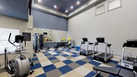 This fitness center offers a comprehensive workout environment designed to support a wide range of exercise routines. The space is immediately striking thanks to its soaring ceilings, which create an open, airy atmosphere that makes the room feel expansive and energizing. The ceiling is finished in a deep charcoal tone, contrasting cleanly with the bright white upper walls, while recessed lighting provides consistent, well-distributed illumination throughout.

The flooring features a bold checkered pattern in blue, beige, and gray tiles, adding a dynamic visual element to the space while providing a durable, easy-to-maintain surface suited for fitness use. A full-length mirror lines one wall, enhancing the sense of depth and allowing users to monitor their form during workouts.

The center is outfitted with multiple treadmills equipped with digital displays, a dumbbell rack with a full range of free weights, a weight bench, and a multi-use strength training station. Stability and medicine balls are also available, supporting core and functional fitness training. The layout is thoughtfully organized, with cardio equipment positioned separately from the free weight and strength training areas, allowing for smooth traffic flow and comfortable use of all equipment simultaneously.