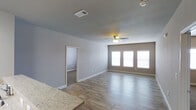 This single-story home in White Settlement offers a clean, modern layout designed for comfortable everyday living. The main living area is a standout feature, showcasing light-toned hardwood-style flooring that runs seamlessly throughout the space, contributing to a bright and cohesive aesthetic. A ceiling fan is centrally positioned overhead, providing both functional air circulation and a subtle decorative touch. A row of windows along the far wall allows generous natural light to flood the room, creating an open and airy atmosphere. Neutral gray walls provide a versatile backdrop that complements a wide range of interior styles. An adjacent room, accessible through a doorway, features carpet flooring, offering a softer, more private retreat — ideal for use as a bedroom. The transition between the two flooring types is clean and well-defined, highlighting thoughtful design choices throughout. Granite-style countertops are visible at the edge of the frame, suggesting a kitchen area nearby that carries the same modern sensibility. HVAC venting integrated into the ceiling ensures year-round climate comfort. Overall, the home presents a well-maintained, move-in-ready environment with consistent finishes and a practical, open feel.