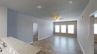This single-story home in White Settlement offers a clean, modern layout designed for comfortable everyday living. The main living area is a standout feature, showcasing light-toned hardwood-style flooring that runs seamlessly throughout the space, contributing to a bright and cohesive aesthetic. A ceiling fan is centrally positioned overhead, providing both functional air circulation and a subtle decorative touch. A row of windows along the far wall allows generous natural light to flood the room, creating an open and airy atmosphere. Neutral gray walls provide a versatile backdrop that complements a wide range of interior styles. An adjacent room, accessible through a doorway, features carpet flooring, offering a softer, more private retreat — ideal for use as a bedroom. The transition between the two flooring types is clean and well-defined, highlighting thoughtful design choices throughout. Granite-style countertops are visible at the edge of the frame, suggesting a kitchen area nearby that carries the same modern sensibility. HVAC venting integrated into the ceiling ensures year-round climate comfort. Overall, the home presents a well-maintained, move-in-ready environment with consistent finishes and a practical, open feel.