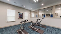 This single-level fitness studio is thoughtfully designed to support a variety of workout disciplines. The main exercise area is anchored by a row of stationary spin bikes positioned on protective floor mats atop a rich, textured blue carpet that adds both visual interest and practicality to the space. Recessed lighting and a ceiling fan ensure the room remains well-lit and comfortable during workouts.

Large floor-to-ceiling mirrors span multiple walls, visually expanding the space and allowing users to monitor their form during exercise. Windows fitted with blinds allow natural light to filter in while maintaining privacy. A framed cycling-themed artwork adds a motivational, sport-specific aesthetic to the room.

An adjacent mirrored section visible through interior windows reveals a dedicated boxing area, complete with a heavy punching bag and a speed bag, offering a separate zone for combat-style training. A wall-mounted flat-screen television is positioned for easy viewing from the cycling area, and a wall-mounted media controller supports guided workout sessions. Hand sanitizer dispensers are also conveniently placed throughout. The overall ambiance is clean, functional, and purpose-built for group or individual fitness activity.