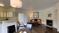 This single-story residence offers an open and well-appointed living area characterized by rich dark hardwood flooring and a warm, neutral color palette on the walls and ceilings. The living room serves as the central gathering space, furnished with a rust-orange sofa accented by patterned throw pillows and complemented by a rectangular glass-top coffee table. A framed piece of wall art adds a decorative focal point, while a tall arc floor lamp provides ambient lighting. The room features smooth, light-toned ceilings with a flush-mount ceiling fixture that enhances the bright, airy atmosphere. Vertical blinds frame a set of sliding glass doors that open to an outdoor area, allowing natural light to filter into the interior and creating a seamless connection between indoor and outdoor living. Adjacent to the living area, a dining space is visible, furnished with sleek dark chairs arranged around a round glass-top table, reinforcing the cohesive contemporary aesthetic. A partial view of what appears to be a kitchen or entryway area with cabinetry is also visible, suggesting a practical and flowing floor plan. Overall, the space presents a clean, comfortable, and functional layout with consistent design elements throughout.