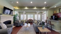 This single-floor community space makes a strong first impression with its grand double-door entry, featuring rich dark wood and glass panels set above a decorative diamond-pattern tile floor. A branded welcome mat hints at the polished, hospitality-driven character of the space. White decorative columns frame the open-plan interior, lending a classic architectural elegance that carries throughout.

The main lounge area is generously proportioned, centered around a striking white stone fireplace with a mounted flat-screen television above the mantle. Comfortable gray upholstered seating, a soft area rug over dark hardwood floors, and lush indoor plants create a warm, inviting atmosphere. Recessed lighting and multiple windows along the rear wall fill the space with natural light and views of the surrounding greenery.

A dedicated dining and meeting area sits just beyond the lounge, furnished with a wooden table and chairs, ideal for gatherings or events. A quieter seating nook with patterned accent chairs provides an additional space for conversation or informal work.

The kitchenette is well-appointed with rich cherry wood cabinetry, a stone tile backsplash, solid-surface countertops, and bar seating — practical for catered events or daily refreshments. Crown molding, soft sage-green walls, and consistent hardwood flooring unify the space with a refined, cohesive aesthetic.