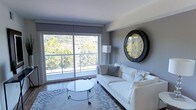 This single-floor space features a thoughtfully designed living area that blends modern aesthetics with a clean, minimalist sensibility. The room is anchored by a large sliding glass door that stretches across much of the exterior wall, allowing abundant natural light to flood the interior and offering views of lush greenery and what appears to be a balcony or terrace beyond. The soft grey walls provide a neutral backdrop that complements the bright, airy atmosphere created by the generous glazing. A plush white sofa serves as the central seating element, styled with geometric patterned cushions that add subtle texture to the monochromatic palette. A sleek, round glass-topped coffee table on casters sits atop a light-toned area rug, contributing to the room's contemporary feel. A standout decorative feature is a large, vintage-style round clock mounted prominently on the grey accent wall, adding character and a focal point to the space. Hardwood flooring runs throughout, grounding the room with warmth. A floor lamp in the corner provides additional ambient lighting. The overall ambiance is calm, bright, and modern.