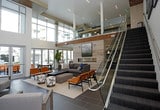 This two-floor residential amenity lounge is thoughtfully designed to offer a comprehensive communal living experience. The space is anchored by a sleek, modern kitchen featuring a large central island with a gas cooktop and integrated range hood, stainless steel appliances including a full-size refrigerator, mosaic tile backsplash, dark wood cabinetry, and pendant lighting — all finished with large-format tile flooring. Adjacent to the kitchen, a dining area with a clean-lined table and seating opens directly onto an outdoor terrace through expansive glass French doors, providing views of the surrounding courtyard and pool area. The lounge level features a warm, inviting seating nook with stone tile and rich wood wall paneling, complemented by a wall-mounted display screen. A dedicated media room offers a large-screen home theater setup with a deep sectional sofa, patterned carpeting with bold orange accents, and built-in shelving. The entertainment zone expands into a generously sized game room equipped with a pool table and shuffleboard table. A glass-railed staircase connects the two floors, with floor-to-ceiling windows flooding the space with natural light and offering views of the outdoor amenity areas below.